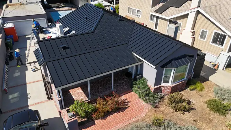 Roofing crew finishing a black standing seam metal roof, with a ladder and materials staged below